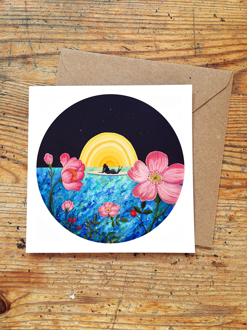 Greeting card with a surfer girl lying on surboard watching a sunset over the sea and flowers on a wooden surface