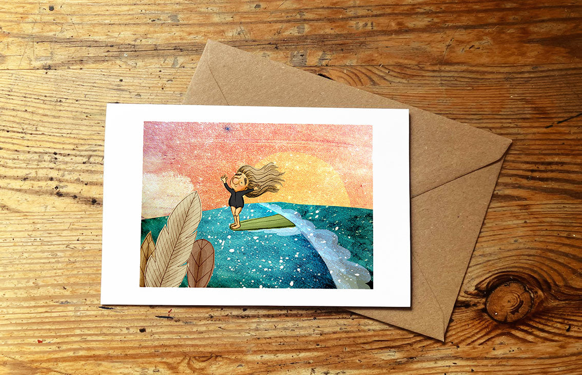 Greeting card with a girl surfing an ocean wave and sunset, with envelope on a wooden surface