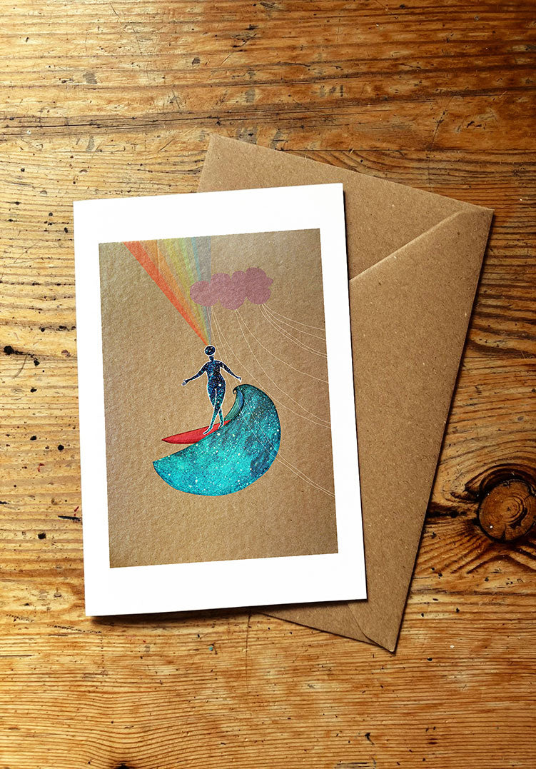 Greeting card with a person on a surfboard riding an ocean wave, rainbow, and cloud design, placed on a wooden surface with a brown envelope.