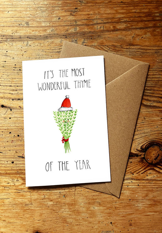 Christmas card with 'It's the most wonderful thyme of the year' text and illustration of a bunch of thyme wearing a santa hat, on a wooden surface