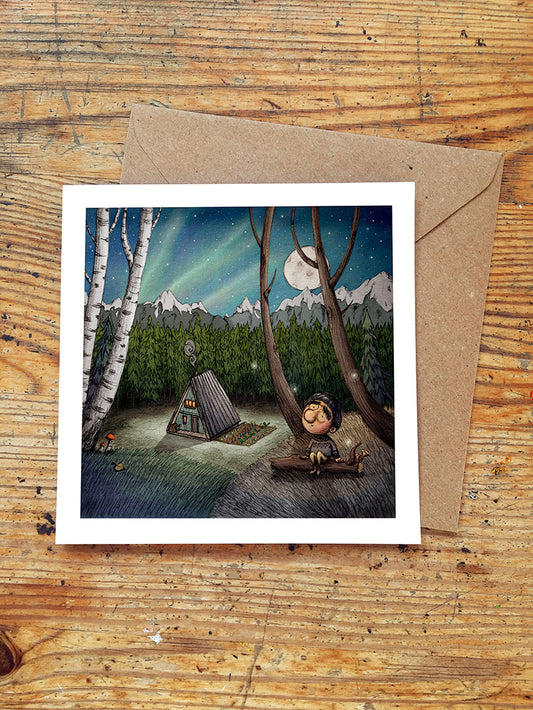 Illustrated card with a mountain and forest scene, cabin, and boy sitting on a log under a starry sky and aurora borealis on a wooden surface.