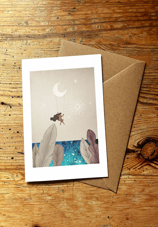 Greeting card with surreal illustration of a woman swinging on a swing hanging from a cresecnt moon above the sea, with an envelope on a wooden surface
