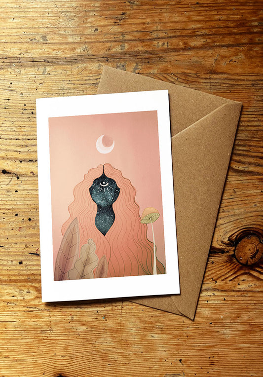 Greeting card with a surreal mystical illustration of a woman with long hair with a crescent moon above and a mushroom and leaves in the foreground on a pink background, placed on a wooden surface with a brown envelope.