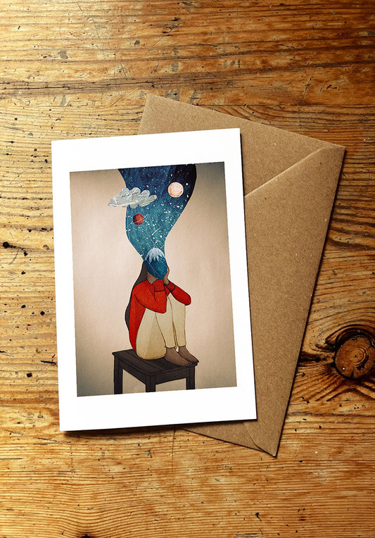Greeting card with a person sitting on a stool and a cosmic design on their head, placed on a wooden surface with a brown envelope.
