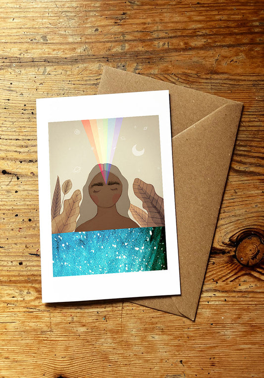 Greeting card with a colorful design of a woman in the sea with a rainbow coming out of her forehead, placed on a wooden surface with a brown envelope.
