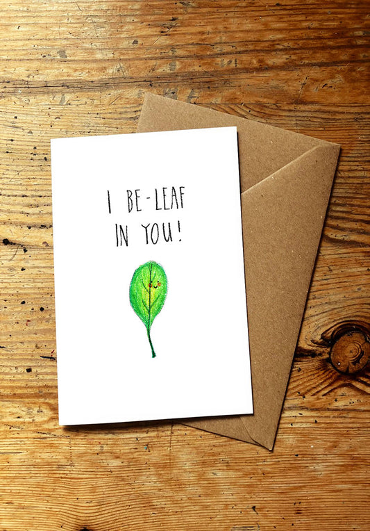 Veg pun greeting card with a smiley leaf illustration and 'I BE-LEAF IN YOU!' text on a wooden surface.