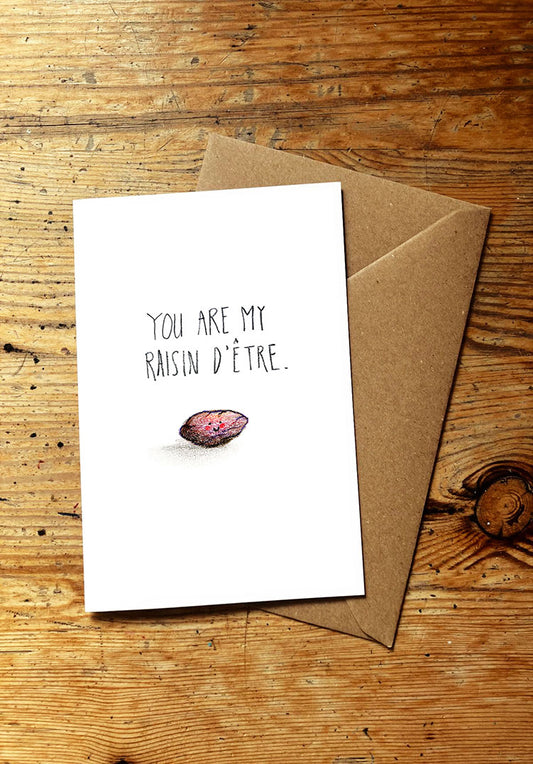 Fruit and veg pun greeting card with 'You are my raisin d'étre' text and a smiling purple raisin on a wooden surface.