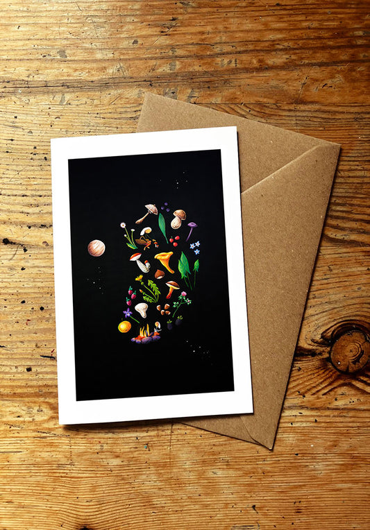 Greeting card with space-themed design featuring planets and stars, and wild edible plants and mushrooms, placed on a wooden surface with a brown envelope.