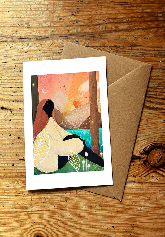 Greeting card with an illustration of a woman sitting in a forest, placed on a wooden surface with a brown envelope.