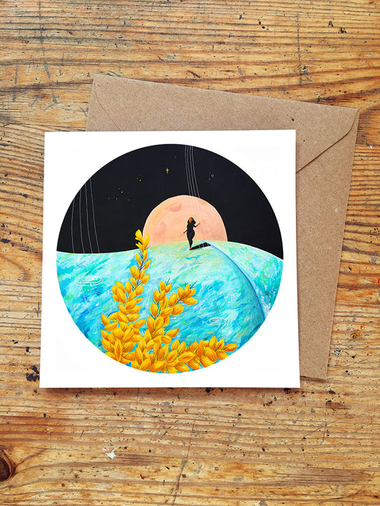 Greeting card with a surf girl riding an ocean wave, full moon rising, yellow gorse flowers in the foreground, placed on a wooden surface with a brown envelope.