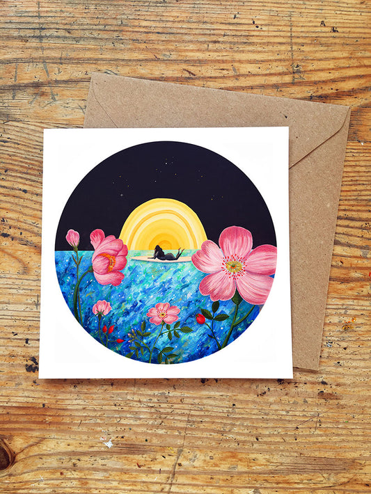 Greeting card with a surfer girl lying on surboard watching a sunset over the sea and flowers on a wooden surface