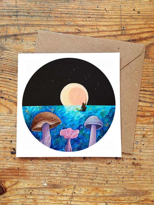 Greeting card with surfer girl waving at a full moon rising over the sea with mushrooms in the foreground and envelope on a wooden surface