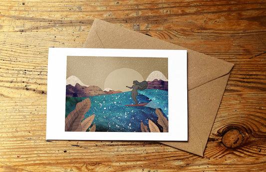 Greeting card with a surf girl and mountain design on a wooden surface with an envelope.