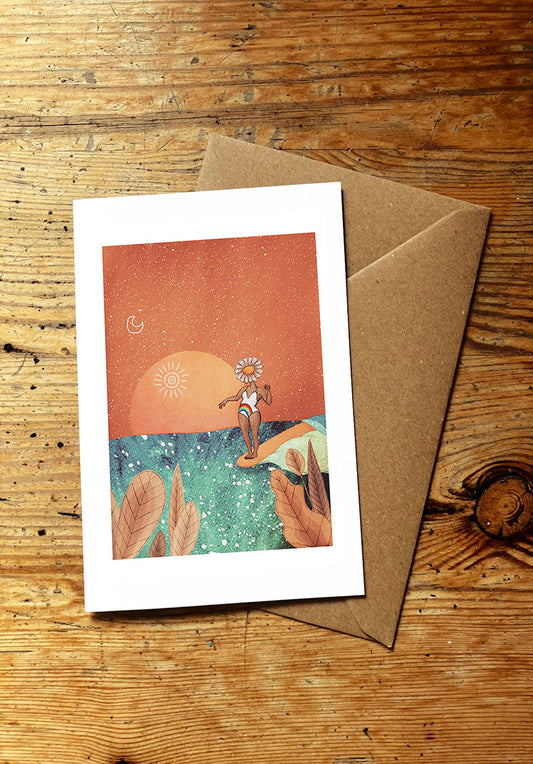 Greeting card with a surf girl surfing an ocean wave and a sunset, placed on a wooden surface with a brown envelope.