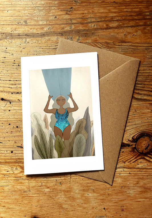 Greeting card with a woman in a blue swimsuit holding a surfboard on her head, with brown envelope on a wooden surface
