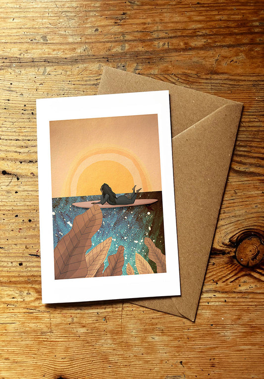 Greeting card with a surf girl lying on a surfboard and sunset design, placed on a wooden surface with a brown envelope.