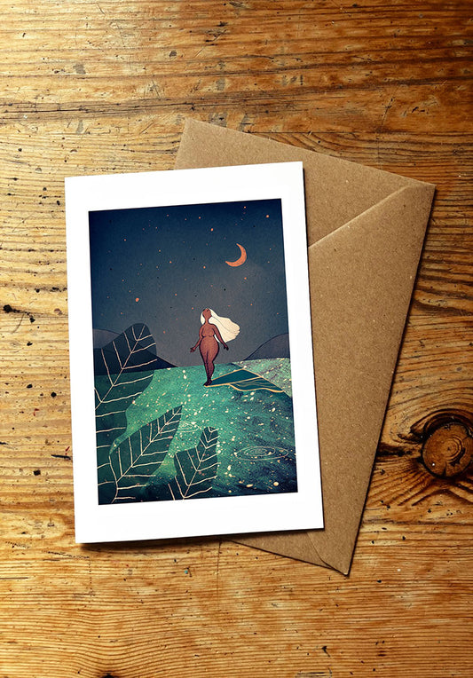 Greeting card with a woman surfing an ocean wave under a night sky with a moon, placed on a wooden surface with a brown envelope.