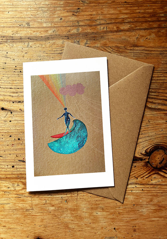 Greeting card with a person on a surfboard riding an ocean wave, rainbow, and cloud design, placed on a wooden surface with a brown envelope.