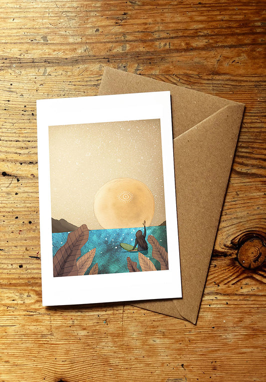Greeting card with a surf girl sitting on a surfboard in the sea and waving at a full moon rising, with brown envelope on a wooden surface