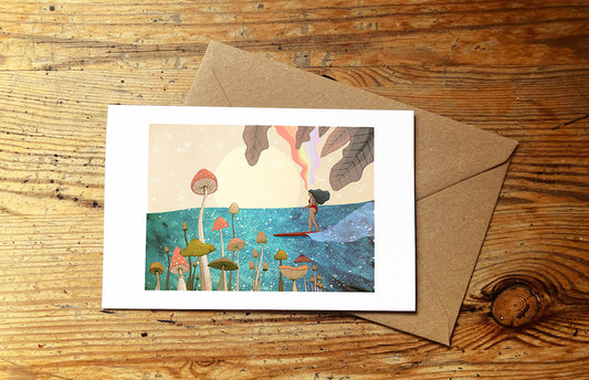 Greeting card with a surf girl riding an ocean wave and mushrooms, and envelope on a wooden surface