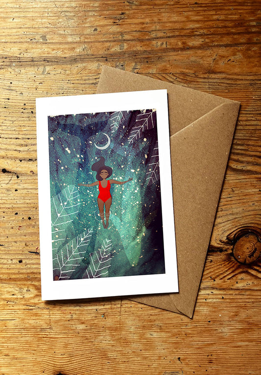 Greeting card with a woman in a red swimsuit floating in water, surrounded by stars and leaves, and two blue whales swimming below her, on a wooden surface with a brown envelope.